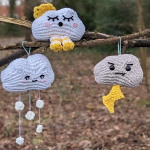 May include: Five crocheted cloud ornaments hanging from tree branches. The clouds have different facial expressions and are decorated with yellow, blue, and white details.