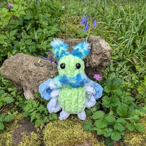May include: A green and blue crocheted plush toy shaped like a butterfly with large black eyes. The toy is sitting on a bed of green moss and surrounded by green plants.