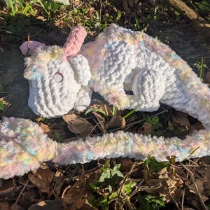 Puede incluir: Un unicornio de crochet blanco con un cuerno rosa y una cola larga multicolor. El unicornio está acostado en un lecho de hojas marrones y plantas verdes.