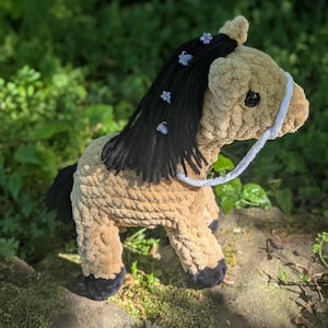 May include: A brown and black crocheted horse toy with a white rope halter. The horse has a black mane and tail with purple flowers woven into the mane.