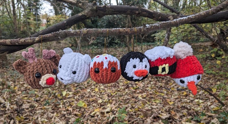 May include: Six crocheted Christmas ornaments hanging from a tree branch. The ornaments are shaped like a reindeer, a polar bear, a gingerbread man, a penguin, Santa Claus, and a snowman.