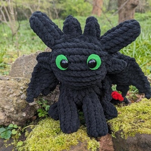以下が含まれることがあります： 緑色の目をした黒い毛糸のぬいぐるみドラゴン。ドラゴンは小さな赤い尻尾を持っており、苔のベッドの上に座っています。