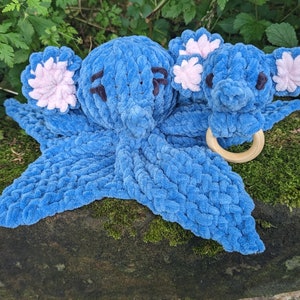 May include: A blue crocheted octopus with pink ears and a wooden teething ring. The octopus is lying on a bed of moss.