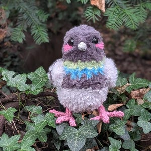 May include: A plush pigeon toy crafted from fuzzy dark gray, light gray, and white yarn. The bird features a vibrant green and blue neck ruff, bright pink stitched cheek patches, black plastic eyes, a small white beak, and pink yarn feet.