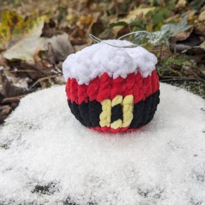 May include: A crocheted Christmas ornament shaped like Santa Claus. The ornament is red, white, and black with a yellow belt buckle. It is sitting on a bed of white faux snow.