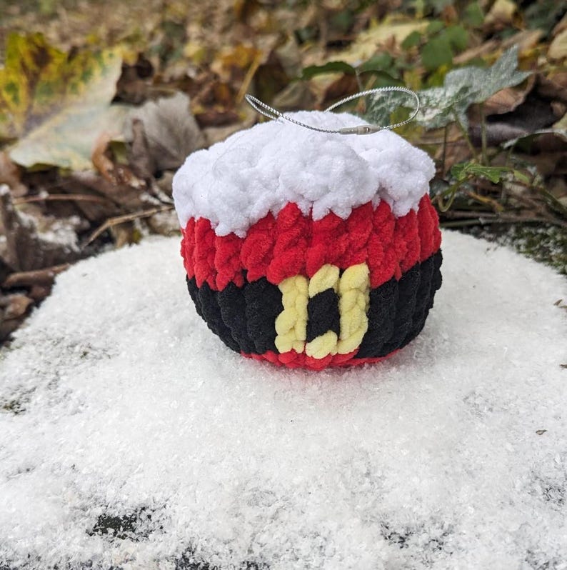 May include: A crocheted Christmas ornament shaped like Santa Claus. The ornament is red, white, and black with a yellow belt buckle. It is sitting on a bed of white faux snow.