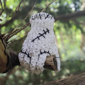 Peut inclure: Main en crochet blanche avec des coutures noires, ressemblant à une main de Frankenstein. La main est suspendue à une branche.