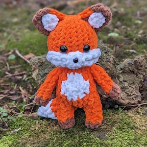 May include: A crocheted orange fox plush toy with white accents. The toy has brown ears and a white belly. The fox is sitting on a bed of green moss.