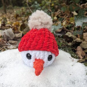 May include: A white crocheted snowman ornament with a red knit hat and a white pom-pom. The snowman has black eyes and an orange carrot nose. The ornament is sitting in a bed of white snow.