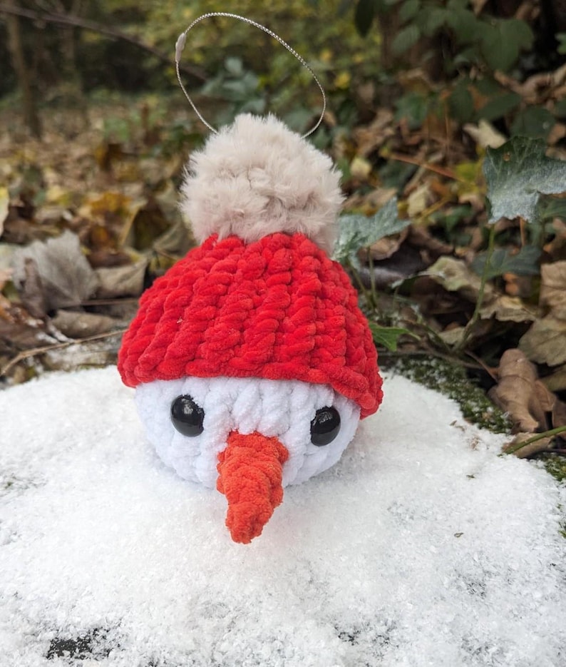 May include: A white crocheted snowman ornament with a red knit hat and a white pom-pom. The snowman has black eyes and an orange carrot nose. The ornament is sitting in a bed of white snow.