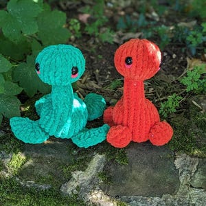 May include: Two crocheted plush toys shaped like dinosaurs. The green dinosaur has black eyes and a pink nose. The red dinosaur has black eyes and a pink nose. The dinosaurs are sitting on a rock covered in moss.