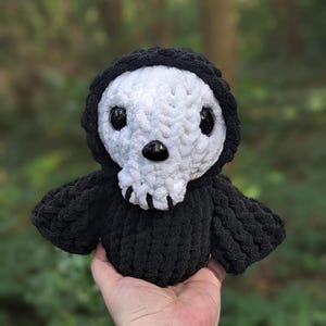 May include: A black and white crocheted plush toy shaped like a skull with large eyes. The toy is wearing a black hooded robe.