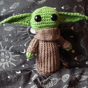 May include: A crocheted green and brown plush toy resembling a character from the Star Wars franchise. The toy has large eyes and a brown knitted sweater.