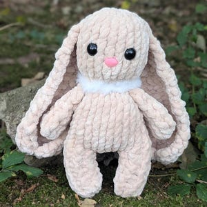 May include: A plush bunny toy in a light beige color, with long floppy ears and a fluffy white collar. The bunny has large black eyes and a small pink nose. The toy is made of a soft, textured material and is sitting on a bed of green moss.