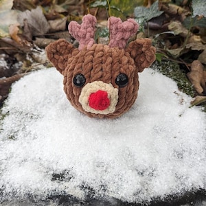 May include: A brown crocheted reindeer ornament with pink antlers and a red nose. The ornament is sitting on a bed of white snow.