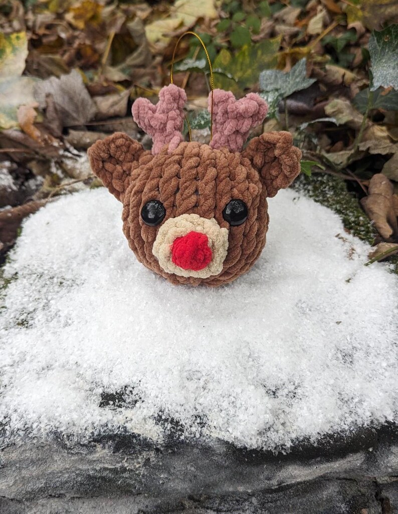 May include: A brown crocheted reindeer ornament with pink antlers and a red nose. The ornament is sitting on a bed of white snow.