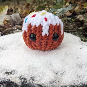 May include: A brown crocheted plush toy shaped like a cake with white frosting and colorful sprinkles. The toy has black eyes and a cute expression.