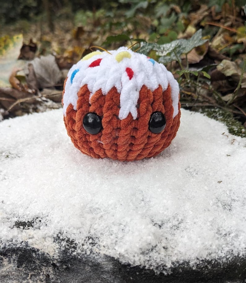 May include: A brown crocheted plush toy shaped like a cake with white frosting and colorful sprinkles. The toy has black eyes and a cute expression.