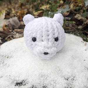 May include: A white crocheted polar bear ornament with black eyes and a black nose. The bear is sitting in a pile of white faux snow.