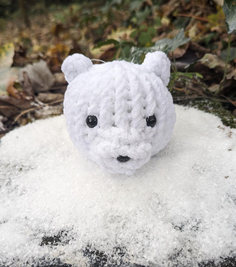 May include: A white crocheted polar bear ornament with black eyes and a black nose. The bear is sitting in a pile of white faux snow.