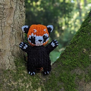 May include: A crocheted red panda plush toy with orange and white fur, black eyes, and black limbs. The toy is sitting on a tree branch covered in moss.