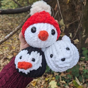May include: Three crocheted plush toys shaped like penguins. The penguin on the left is black and white with a red beak. The penguin in the middle is white with a red hat and a red beak. The penguin on the right is white with a black beak.