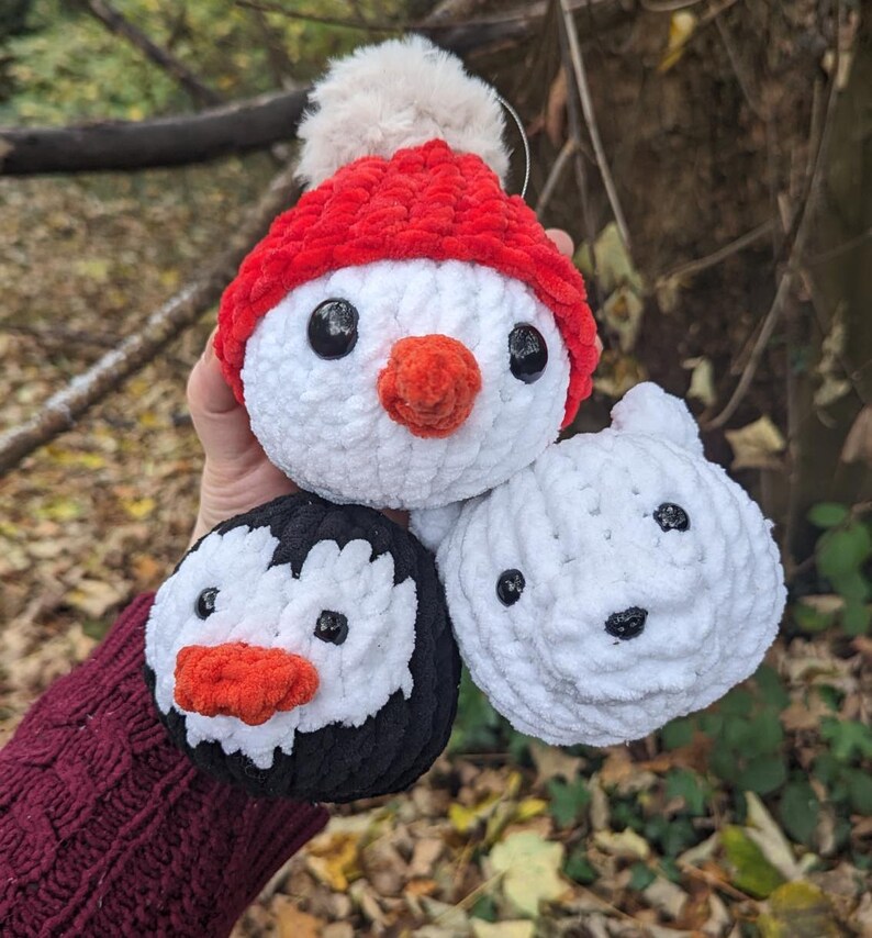 May include: Three crocheted plush toys shaped like penguins. The penguin on the left is black and white with a red beak. The penguin in the middle is white with a red hat and a red beak. The penguin on the right is white with a black beak.