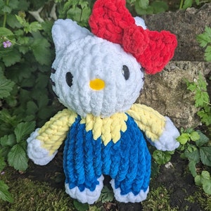 May include: A white crocheted plush toy shaped like a cat with a red bow, blue and yellow clothing, and a yellow nose.