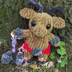 Op de afbeelding: Een bruin gebreid pluche speelgoed in de vorm van een geit met een rood-zwarte riem. Het speelgoed heeft een zilveren neusring en zwarte wollen hoorns.