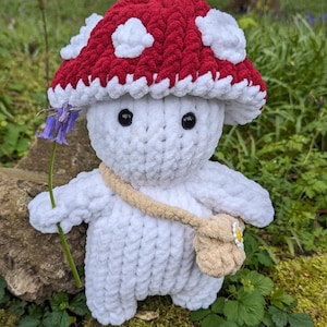 May include: A white crocheted mushroom-shaped plush toy with a red and white crocheted hat. The toy is holding a green stem with a purple flower. The toy is sitting on a bed of green moss.