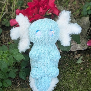 May include: A light blue crocheted axolotl plush toy with white fluffy gills and accents. The toy has two clear, iridescent eyes and is set against a backdrop of red roses and greenery. The plush is soft and cuddly.