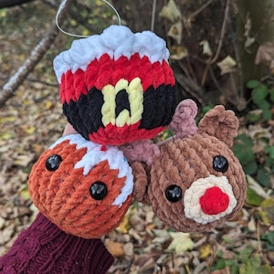 Puede incluir: Tres adornos de crochet, un Papá Noel, un hombre de jengibre y un reno. Los adornos están hechos con un hilo peludo y tienen ojos negros. El adorno de Papá Noel tiene una barba blanca y un cinturón negro.