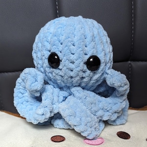 May include: A light blue crocheted octopus plush toy with black button eyes. The octopus is sitting on a white surface with brown and pink accents.