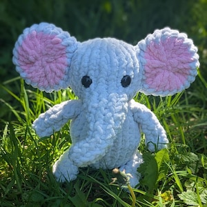 May include: A handmade, light blue crochet elephant plush toy with pink inner ears and black safety eyes. The elephant is sitting in green grass, showcasing its soft, textured yarn construction. A charming, cuddly stuffed animal.