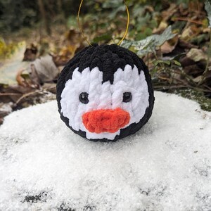 May include: A black and white crocheted penguin ornament with a red beak and black eyes. The ornament is hanging from a gold string.