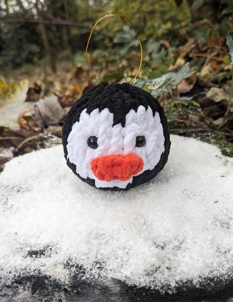 May include: A black and white crocheted penguin ornament with a red beak and black eyes. The ornament is hanging from a gold string.