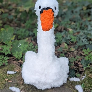 May include: A fluffy white swan toy with an orange beak and black eyes. The soft toy is positioned on a moss-covered surface. Several white feathers are scattered around the base of the swan.