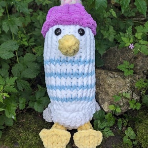 May include: A white crocheted penguin wearing a pink knit hat with a multi-colored pom-pom. The penguin has a yellow beak and yellow feet.