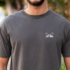 May include: A dark gray t-shirt with a white cat ear design and the text "CAT DAD" on the front.