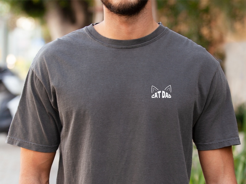 May include: A dark gray t-shirt with a white cat ear design and the text "CAT DAD" on the front.