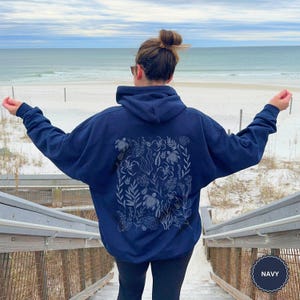 May include: Navy blue hooded sweatshirt with a detailed design on the back featuring sea turtles and aquatic plants. The sweatshirt is worn by a person on wooden steps leading to a beach with the sea in the background.