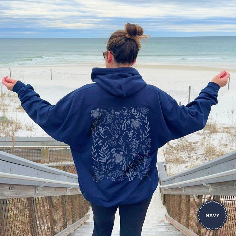 May include: Navy blue hooded sweatshirt with a detailed design on the back featuring sea turtles and aquatic plants. The sweatshirt is worn by a person on wooden steps leading to a beach with the sea in the background.