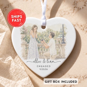 May include: A white heart-shaped ceramic ornament featuring a watercolor illustration of a proposal scene. The text reads "ellie & liam", "ENGAGED", and "9/20/26". A white ribbon is attached to the top for hanging, and the design includes a park setting.