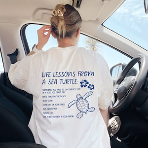 Könnte beinhalten: Weißes T-Shirt mit blauem Text, der "Life Lessons From A Sea Turtle:" lautet und eine Liste von Lebenslektionen enthält. Der Text wird von einer blauen Illustration einer Meeresschildkröte gefolgt.