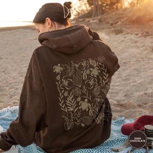 May include: Dark chocolate brown hoodie featuring a floral and turtle design on the back. The hoodie has a drawstring hood and a kangaroo pocket. The design includes turtles, flowers, and leaves in a light beige colour. The hoodie is worn on a beach.
