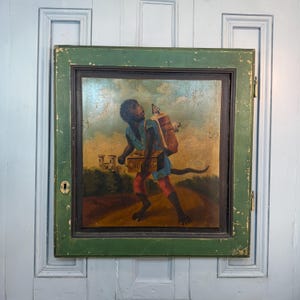 Op de afbeelding: Oude, ingelijste schildering met een aapachtig figuur dat een grote container en een kist draagt. De figuur draagt blauwe en rode kleding, tegen een landschap met een kasteel. De lijst is groen en vertoont slijtage.