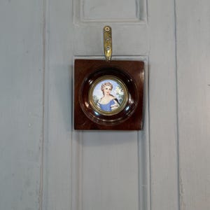 May include: A small, framed portrait of a woman in a blue dress with a white collar. The portrait is set against a light blue background and is framed in a dark wood frame.