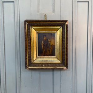 May include: A framed oil painting of a young boy in a brown vest and blue pants standing in front of a stone fountain. The painting is set in a dark brown wooden frame with gold accents.