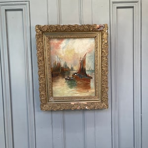 Puede incluir: Una pintura al óleo enmarcada que representa un paisaje marítimo con varios veleros en un puerto. El cielo es de un rosa y naranja suave, y el agua es de un azul tranquilo. La pintura está en un marco dorado con detalles ornamentados.