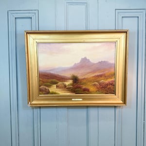 Könnte beinhalten: Ein gerahmtes Landschaftsgemälde mit einem goldfarbenen Rahmen. Das Gemälde zeigt eine Bergkette, einen Bach und farbenfrohes Laub in Braun-, Grün- und Lilatönen. Der Himmel ist ein zartes Rosa und Gelb. Das Gemälde ist an einer hellblauen Tür befestigt.